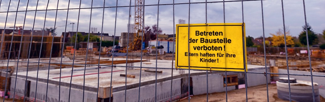Das Bild zeigt eine Baustelle, welche mit einem Zaun abgesperrt ist. An dem Zaun hängt ein gelbes Warnschild, auf dem "Betreten der Baustelle verboten! Eltern haften für ihre Kinder!" steht. 