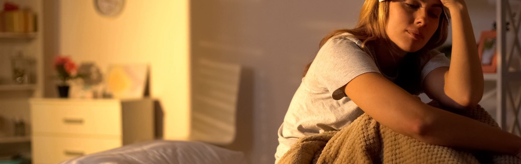 Eine Frau sitzt krank und erschöpft in ihrem Bett. Sie hält sich ein Wärmekissen an die Stirn. 
