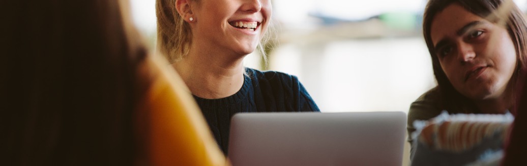 Junge Studentin lacht am Tisch mit ihren Kommilitonen während sie an ihrem Laptop arbeitet.