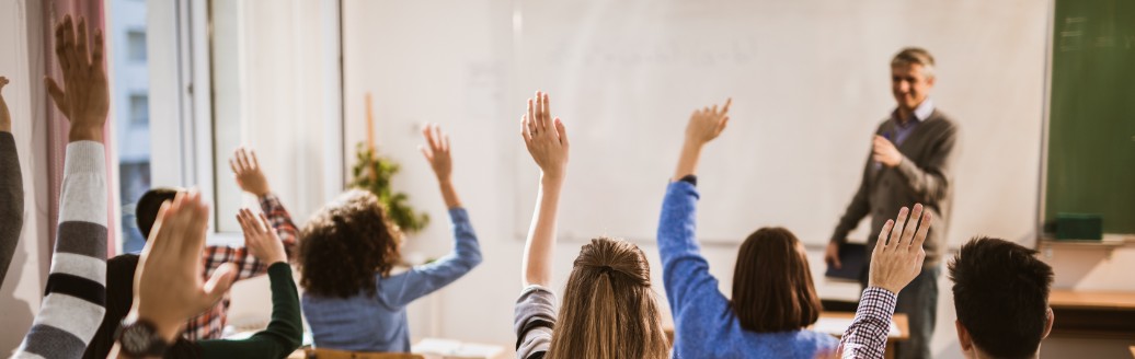 Ein Lehrer steht vor eine Klasse in einem Klassenzimmer. Viele Schüler melden sich. 