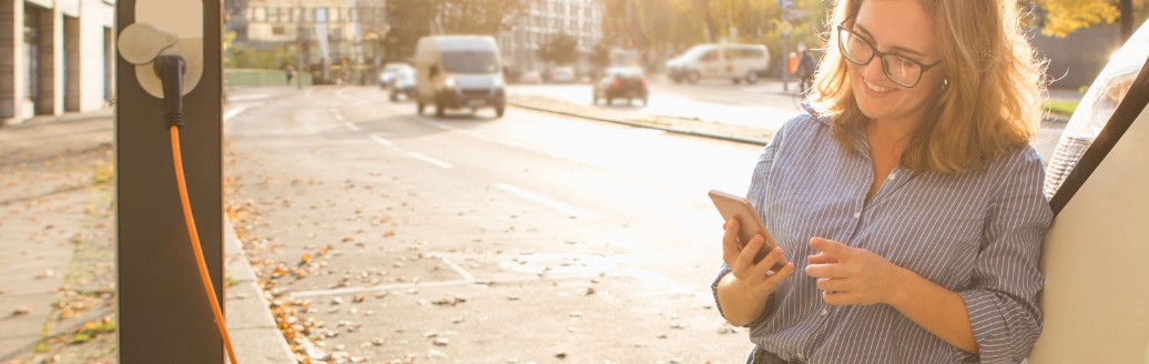 Eine Frau lehnt sich an ihr Auto, welches gerade auflädt, und schaut auf ihr Smartphone.