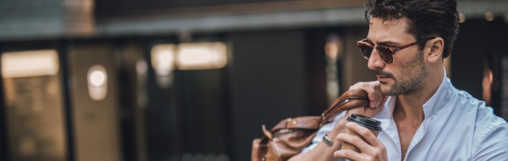 Junger Mann mit Sonnenbrille trinkt einen Kaffee während er seine Reisetasche über eine Schulter sowie eine Luxusuhr trägt. 