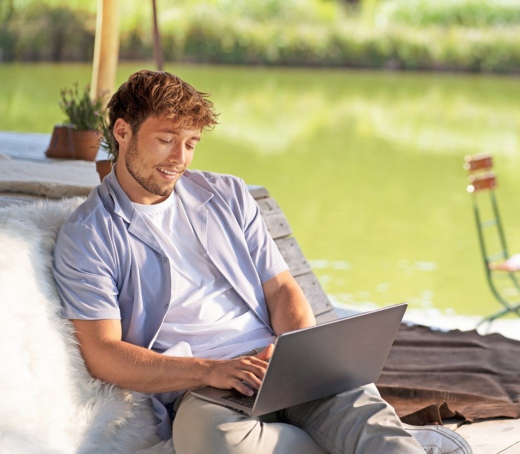 Junger Mann mit T-Shirt und Sommerhemd sitzt an einem See und schaut in ein Laptop