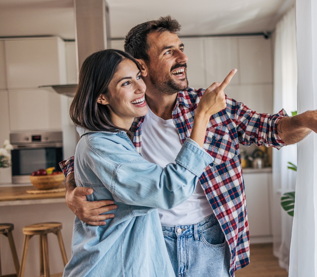 Ein junges Pärchen steht gemeinsam in einer Wohnung und schaut lachend aus dem Fenster.