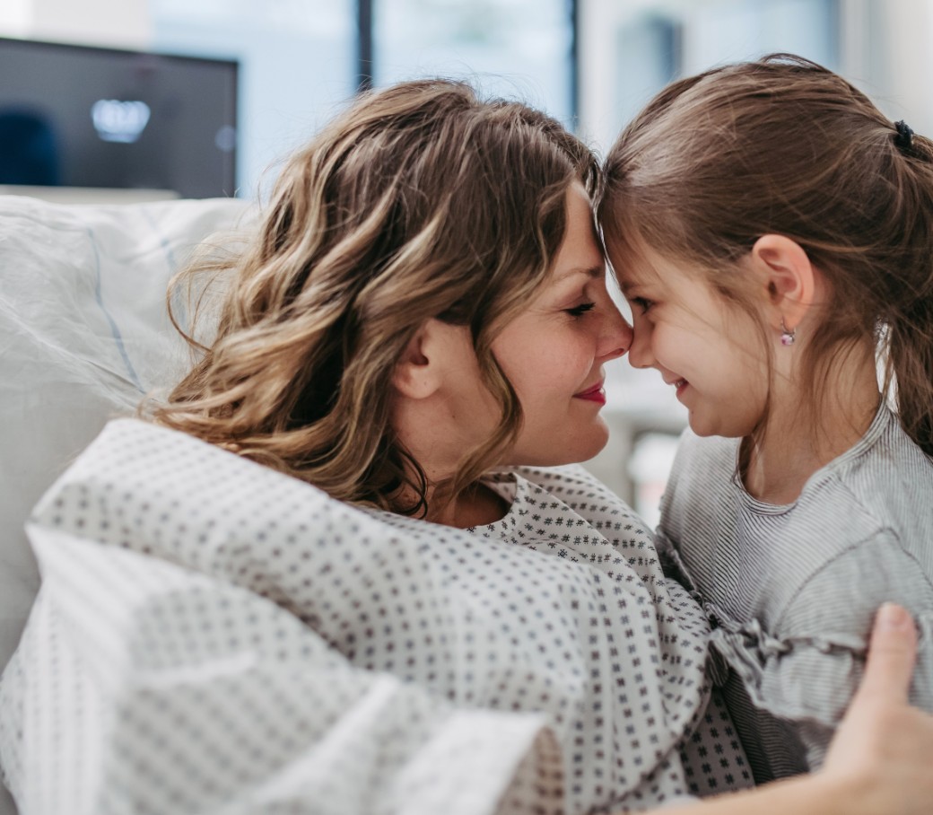Eine Frau im Krankenbett und ihre kleine Tochter geben sich einen Nasenkuss