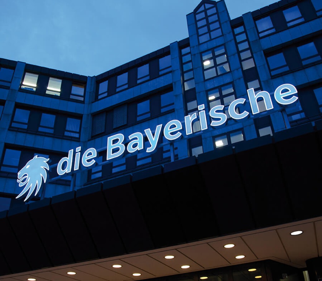 Eingang eines Bürogebäudes mit dem beleuchteten Schriftzug "die Bayerische" bei Dämmerung.