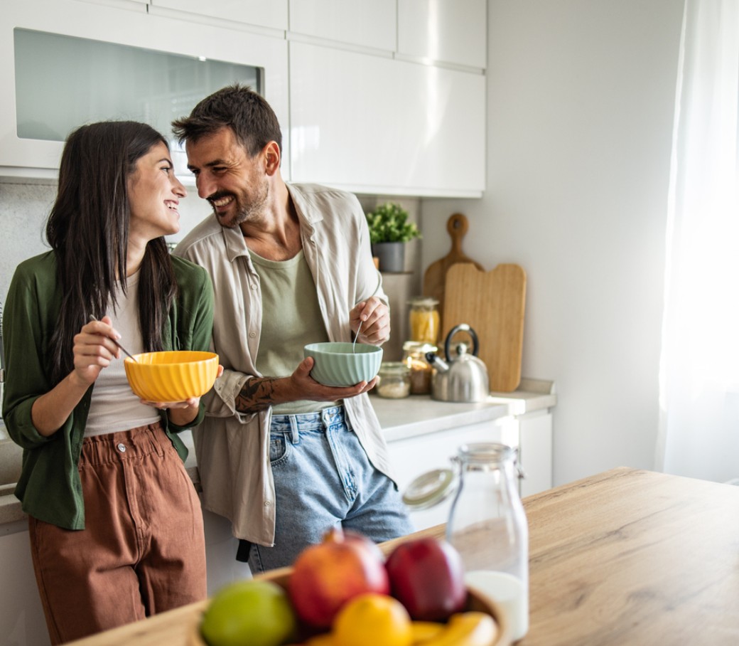 Ein junges Paar steht mit Müslischalen in der Hand steht lachend in der Küche