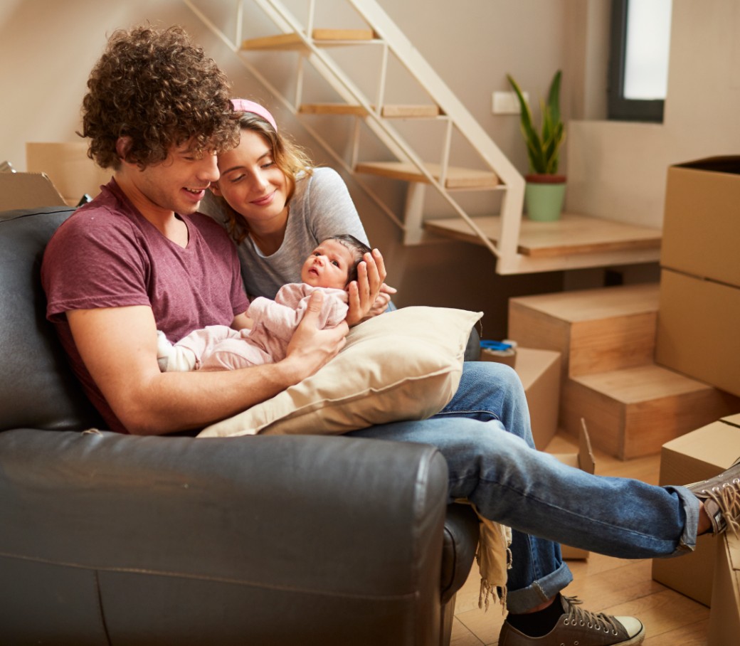 Junges Pärchen mit Baby im Arm sitzen im Wohnzimmer, voll mit Umzugskartons