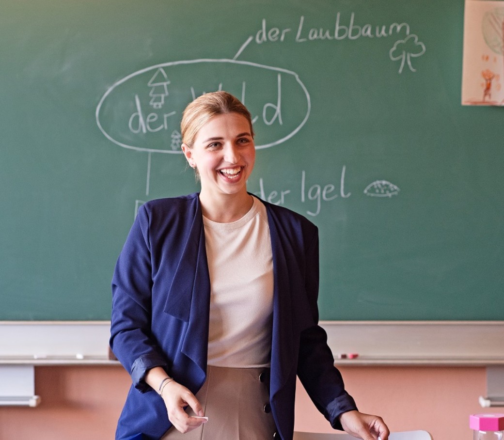 Eine junge Lehrerin steht mit dem Rücken zur Tafel und lächelt