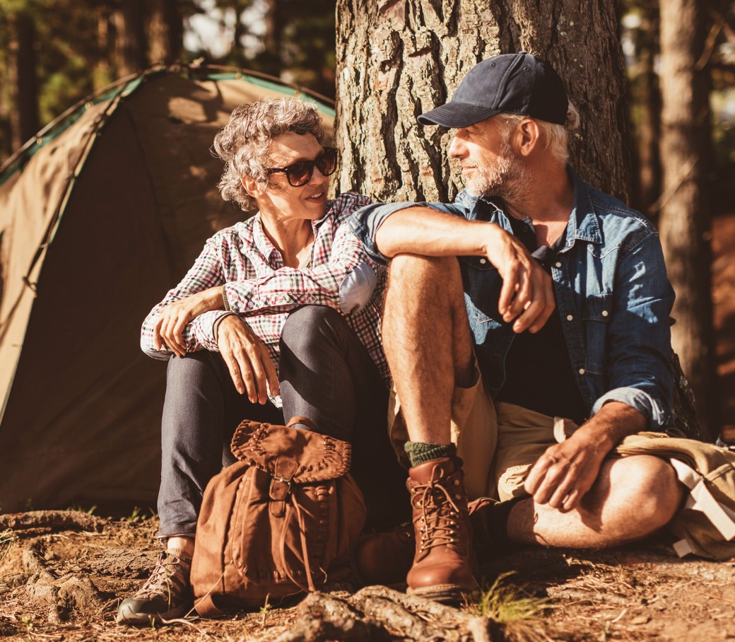 Eine Frau ein Mann in Wanderkleidung sitzen nebeneinander angelehnt an einen Baum und unterhalten sich.