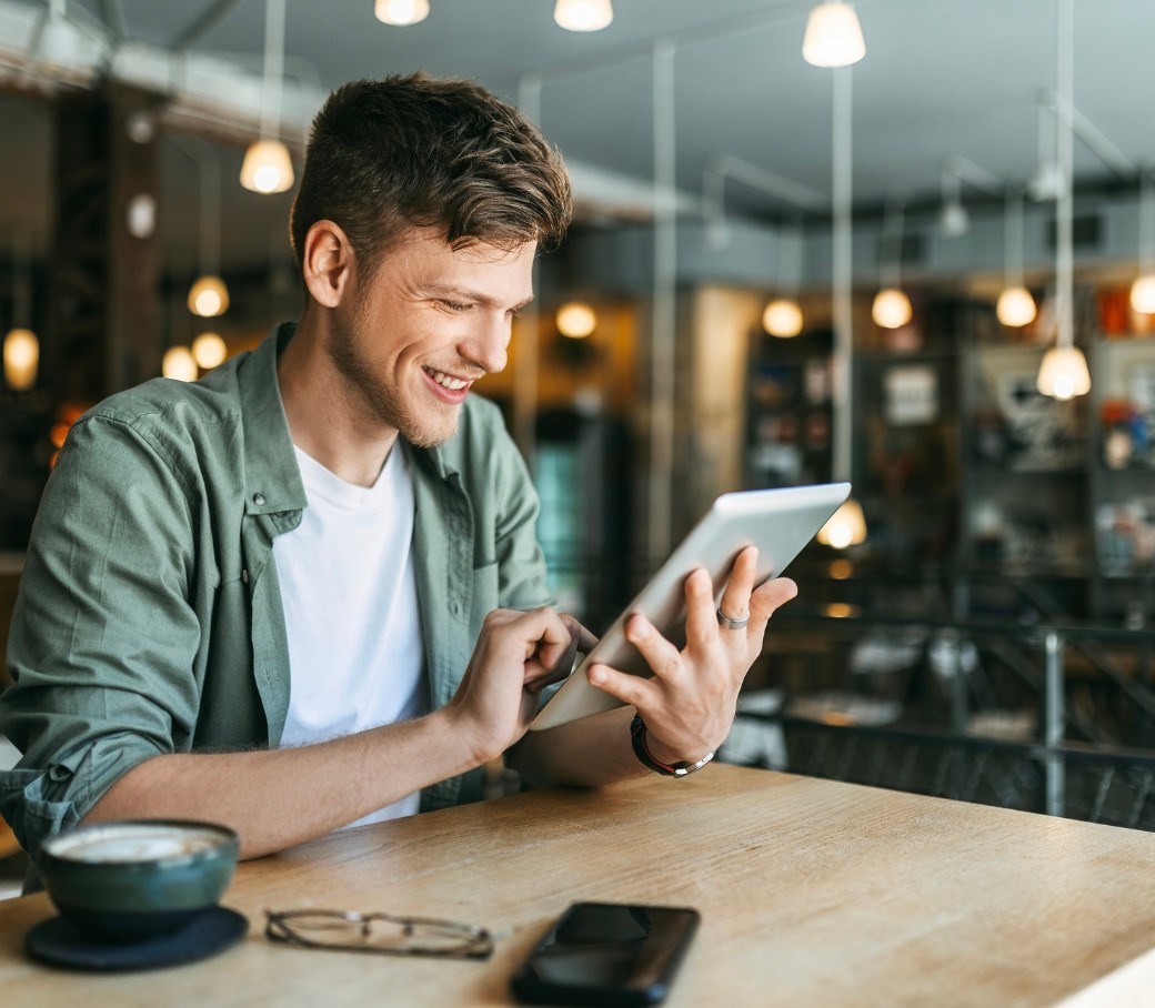 Junger Mann mit grünem Hemd liest in einem Bücher-Cafe auf einem Tablet und lächelt