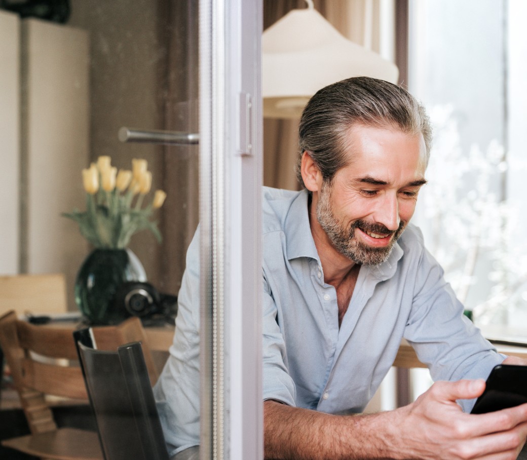 Ein Mann sitzt am offenen Fenster und lächelt in sein Mobiltelefon