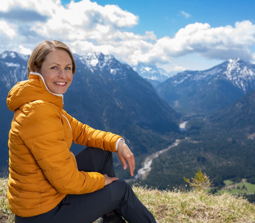 Magdalena Neuner sitzt auf einer Wiese in den Bergen und blickt in die Kamera.
