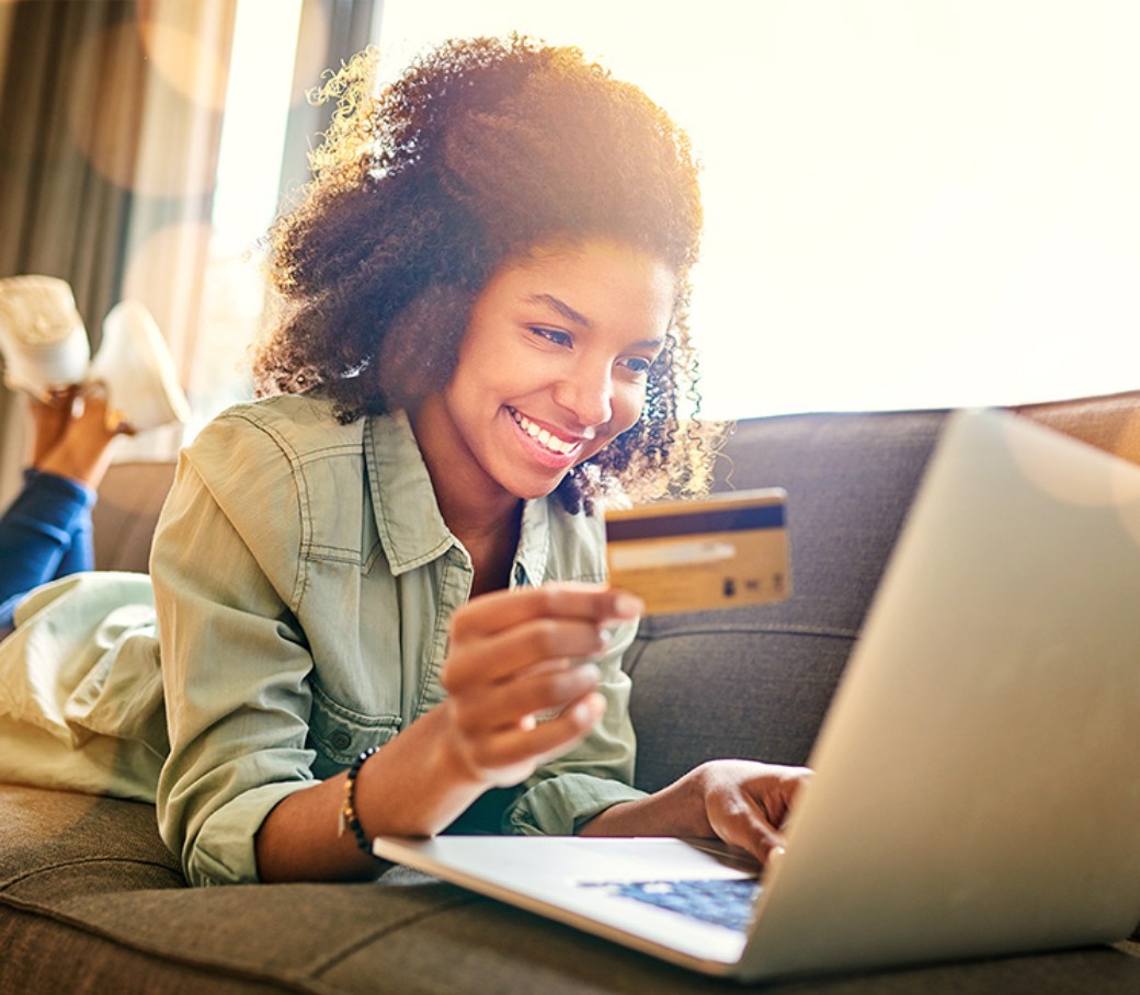 Eine junge Frau liegt auf dem Sofa und tippt lächelnd die Daten ihrer Kreditkarte in ihren Laptop ein.