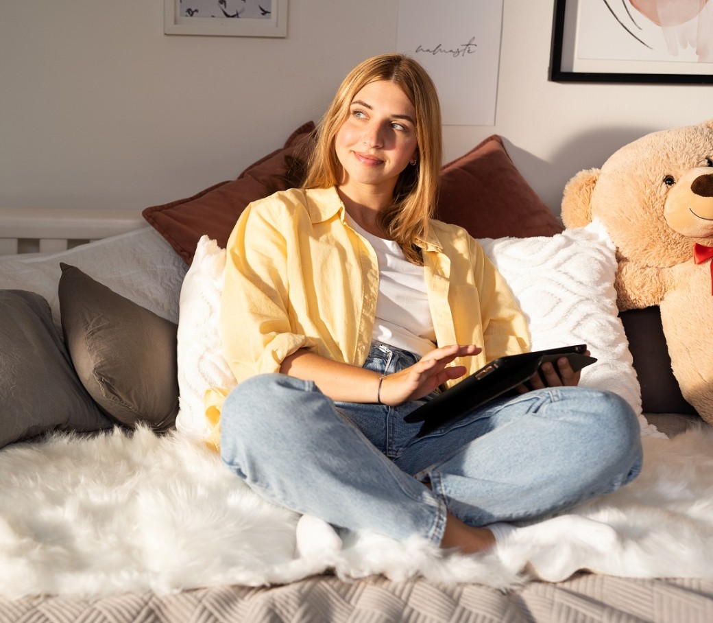 Eine junge Frau sitzt mit einem Riesen-Teddy auf ihrem Bett und hält ein Tablet in der Hand.