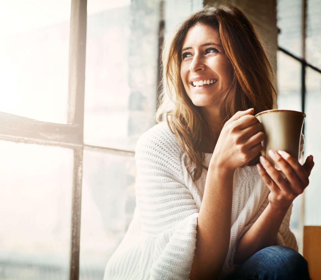 Eine lächelnde Frau sitzt am Fenster mit einer Tasse in der Hand.