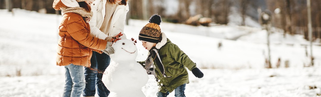 Eine Mutter baut mit ihren zwei Kindern einen Schneemann in einer Winterlandschaft. 