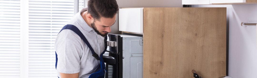 Ein Elektriker steht in einer Küche und untersucht einen Ofen, den er zuvor leicht aus dem Küchenschrank gezogen hat.