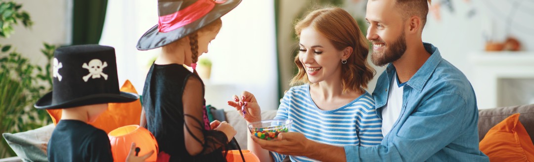 Zwei auf dem Sofa sitzende Eltern geben ihren verkleideten Kindern Süßigkeiten in ihren Halloween-Eimer. 