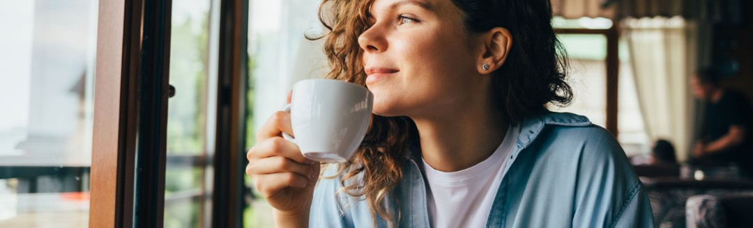 Eine junge Frau schaut aus dem Fenster, während sie einen Kaffee trinkt. 