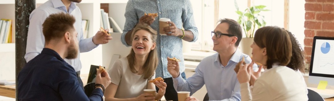 Eine Gruppe von Kollegen sitzt lachend zusammen im Büro an einem Tisch und isst Pizza. 