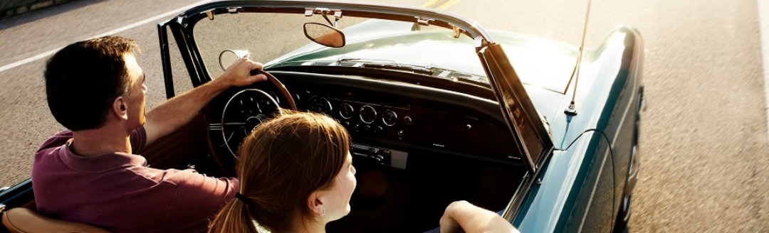 Eine Frau und ein Mann fahren zusammen in einem Oldtimer über eine Landstraße.