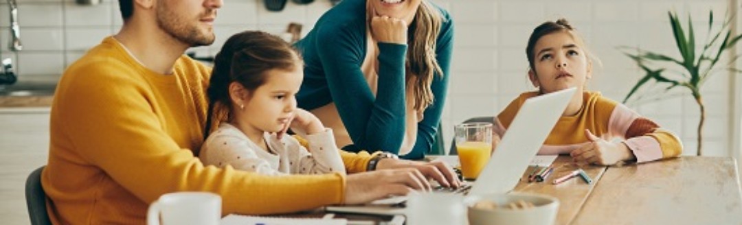 Eine junge Familie sitzt zusammen am Küchentisch. Der Vater arbeitet an seinem Laptop.