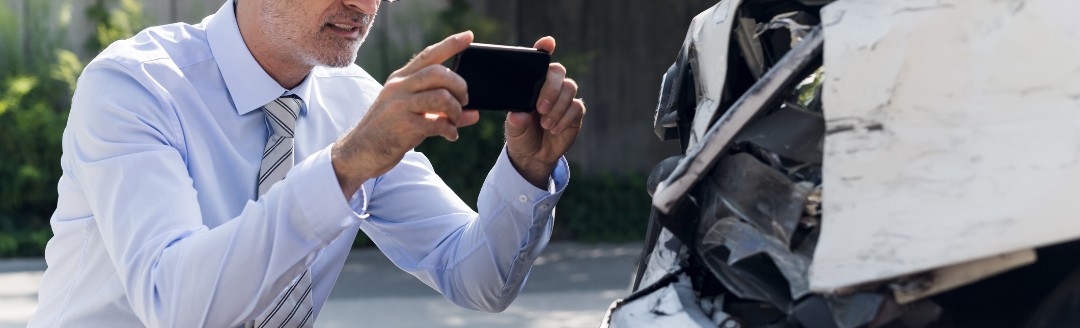 Ein älterer Mann fotografiert mit seinem Mobiltelefon sein Auto, dass einen Schaden erlitten hat.