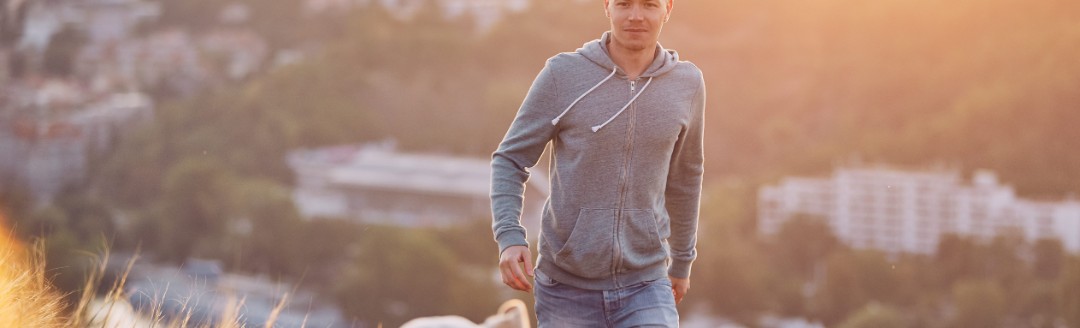 Ein Mann spaziert mit seinem Hund auf einer Wiese spazieren. Im Hintergrund ist eine Stadt zu sehen. 