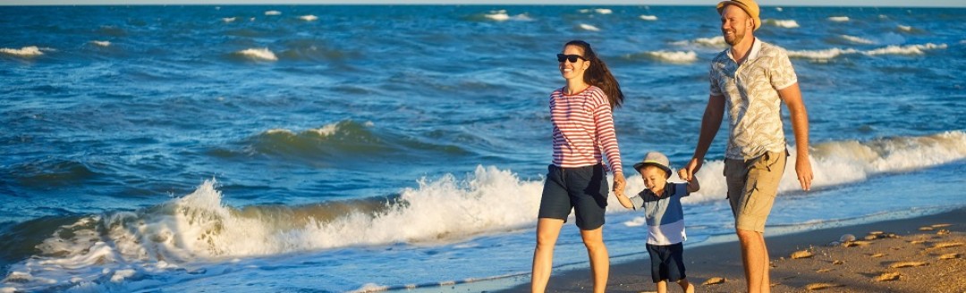 Ein Pärchen läuft mit ihrem Kleinkind Hand in Hand einen Strand entlang. 