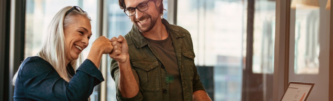 Ein junger Mann und eine junge Frau sitzen zusammen am Laptop im Büro und geben sich die Faust.