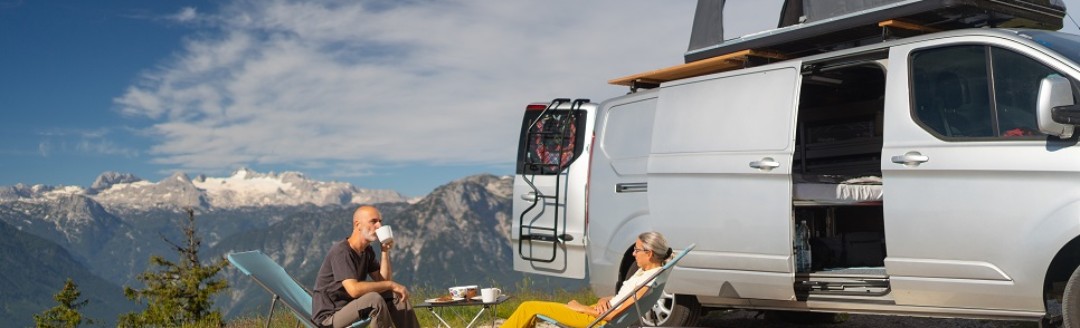 Eine Frau und ein Mann sitzen zusammen in einem Liegestuhl vor ihrem Campervan in den Bergen. Vor ihnen stehen zwei Solarpanels.