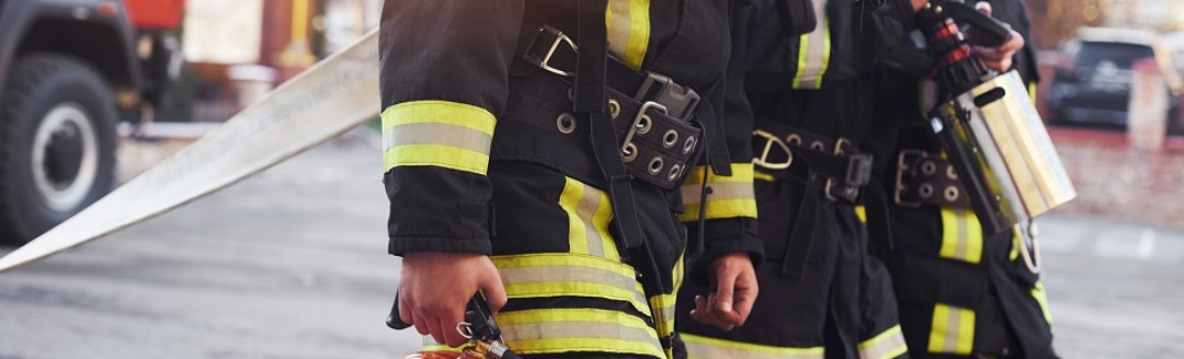 Drei Feuerwehrleute laufen mit Equipment herum. Ihre Uniformen und Equipment stehen im Fokus des Bildes. 