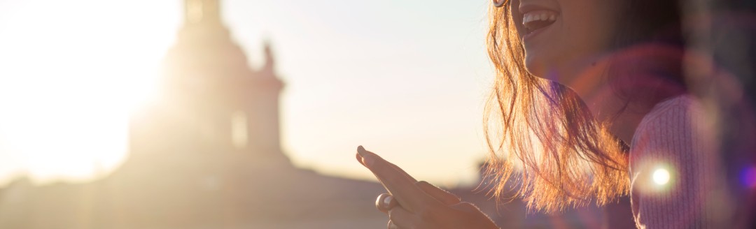 Eine junge Frau schaut lächelnd von ihrem Smartphone hoch, während sie vor einem größeren, weißen Gebäude sitzt. 