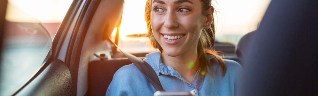 Frau sitzt auf der Rückbank, schaut lachend aus dem Fenster hinaus während sie ein Telefon in der Hand hält.