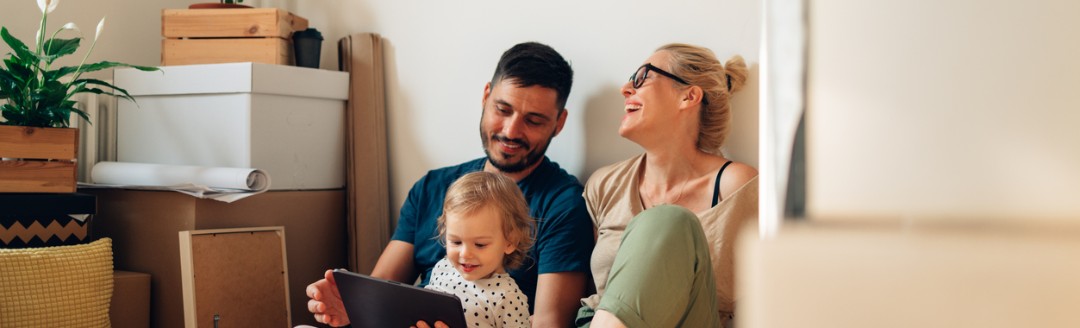 Eine Familie sitzt zwischen Umzugskartons auf dem Boden; ein Mann und ein kleines Kind schauen gemeinsam auf ein Tablet, während eine Frau daneben lachend zusieht.