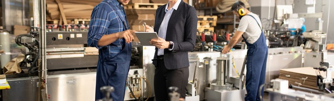 Eine Business-Frau und ein Bauarbeiter sprechen zusammen über ein Dokument, welches die Frau in der Hand hält. Die Umgebung ist eine Fabrik. 