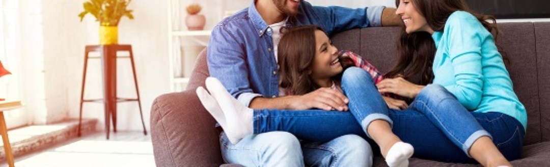 Zwei junge Eltern sitzen lachend mit ihrer Tochter auf dem Sofa.