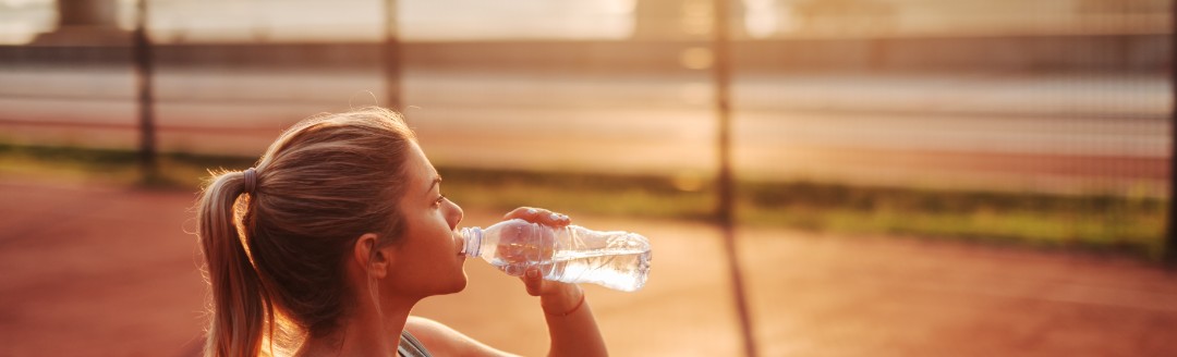 Eine junge Frau steht in einem kurzen Top auf einem Basketballfeld und trinkt Wasser aus einer Flasche.