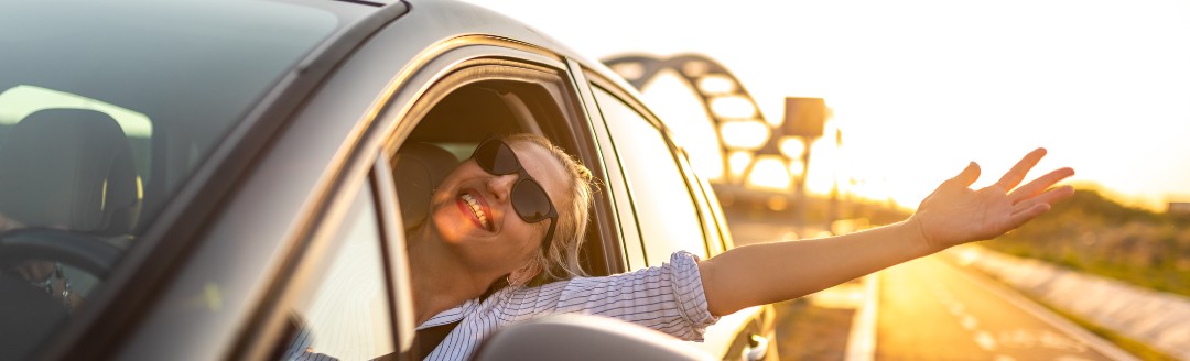 Eine junge blonde Frau fährt in glücklich in der Sonne ihr Auto, trägt eine Sonnenbrille, streckt einen Arm aus dem Fahrerfenster und lacht.