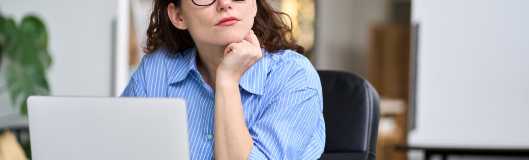 Nachdenkliche Frau mit Brille sitzt am Schreibtisch vor einem Laptop, stützt ihr Kinn auf die Hand und schaut zur Seite.