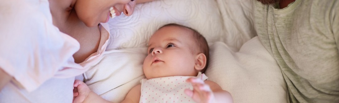 Eine Frau und ein Mann liegen gemeinsam im Bett. In der Mitte ihr Neugeborenes, welches sie verliebt anschauen. 
