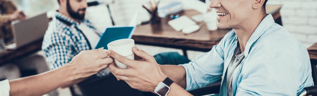 Ein junger Mann sitzt im Rollstuhl und ihm wird eine Tasse Kaffee gereicht. Mit ihm sitzt ein Kollege im Büro. 