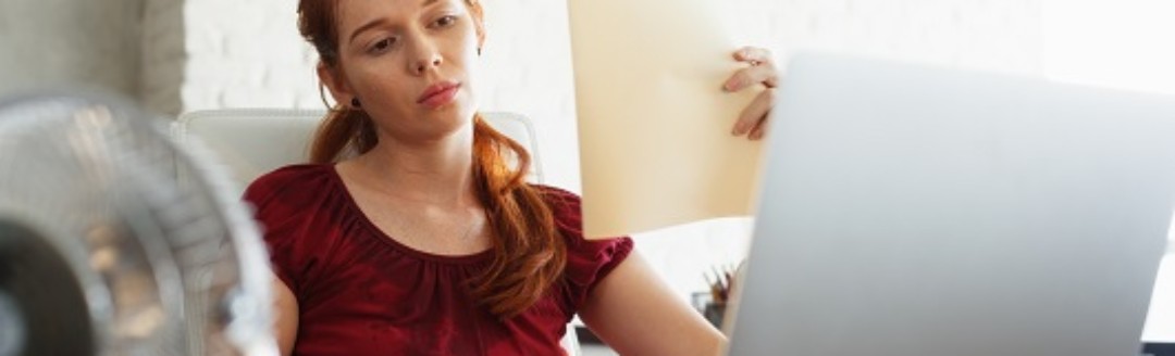 Eine Frau sitzt verschwitzt am Laptop, vor ihr ein Ventilator und sie fächert sich Luft mit Unterlagen zu. 