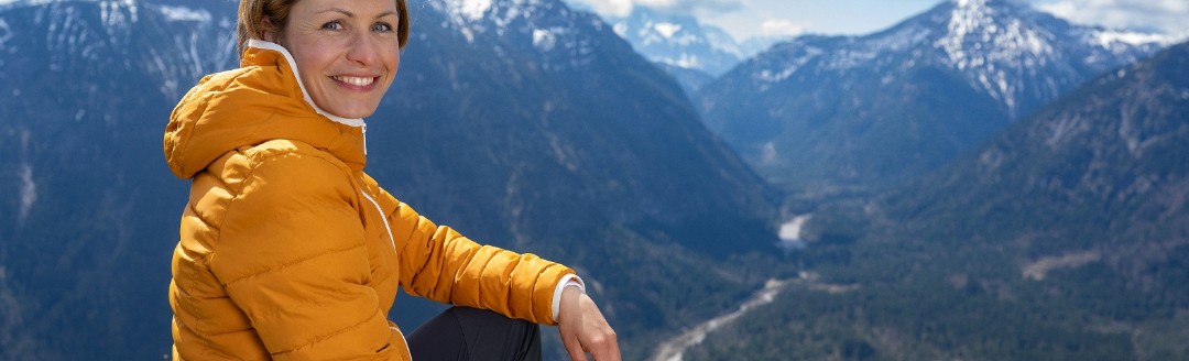 Magdalena Neuner sitzt auf einer Wiese in den Bergen und blickt in die Kamera.