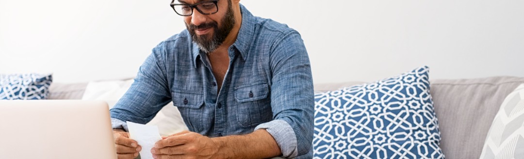Ein Mann sitzt auf einem Sofa, vor ihm steht auf dem Wohnzimmertisch ein Laptop. Der Mann hält einen Kassenzettel in der Hand. 