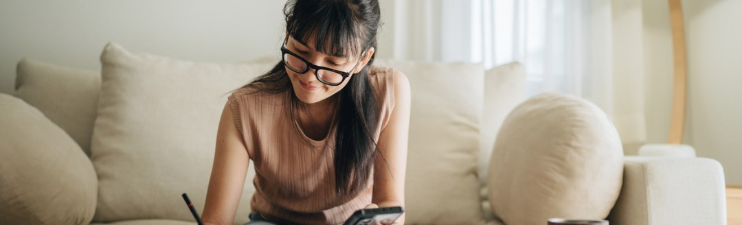 Eine junge Frau sitzt auf einem Sofa, schaut auf Unterlagen auf dem Tisch und macht Notizen, während sie ein Smartphone in der Hand hält.