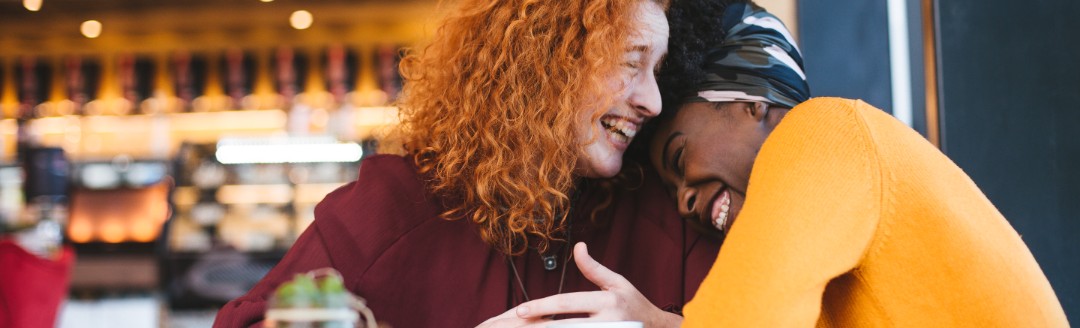 Zwei Frauen sitzen in einem Café und liegen sich lachend in den Armen. 