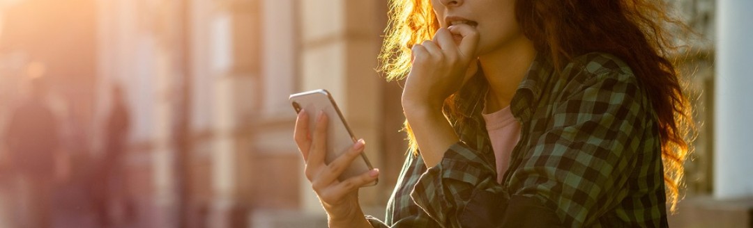 Eine junge Frau sitzt draußen auf einer Steinstufe und schaut gespannt in ihr Smartphone. 