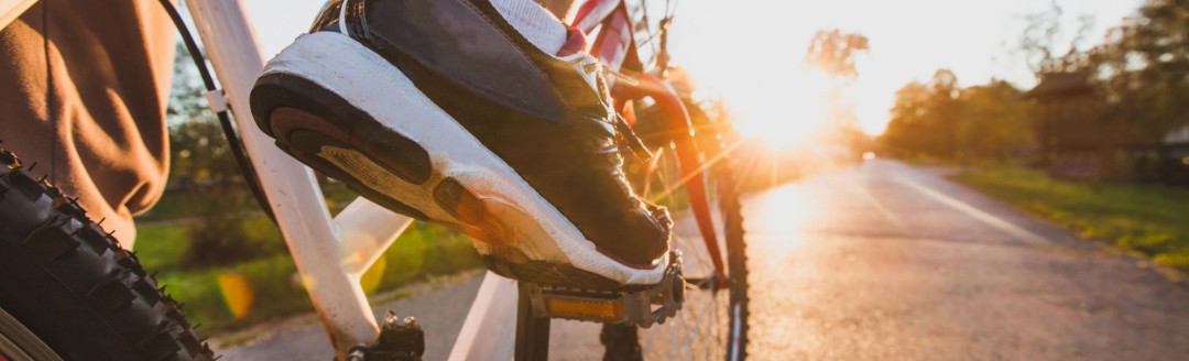 Ein Mann fährt sein Fahrrad im Sonnenuntergang auf einem asphaltierten Weg. Im Fokus stehen seine Sportschuhe.